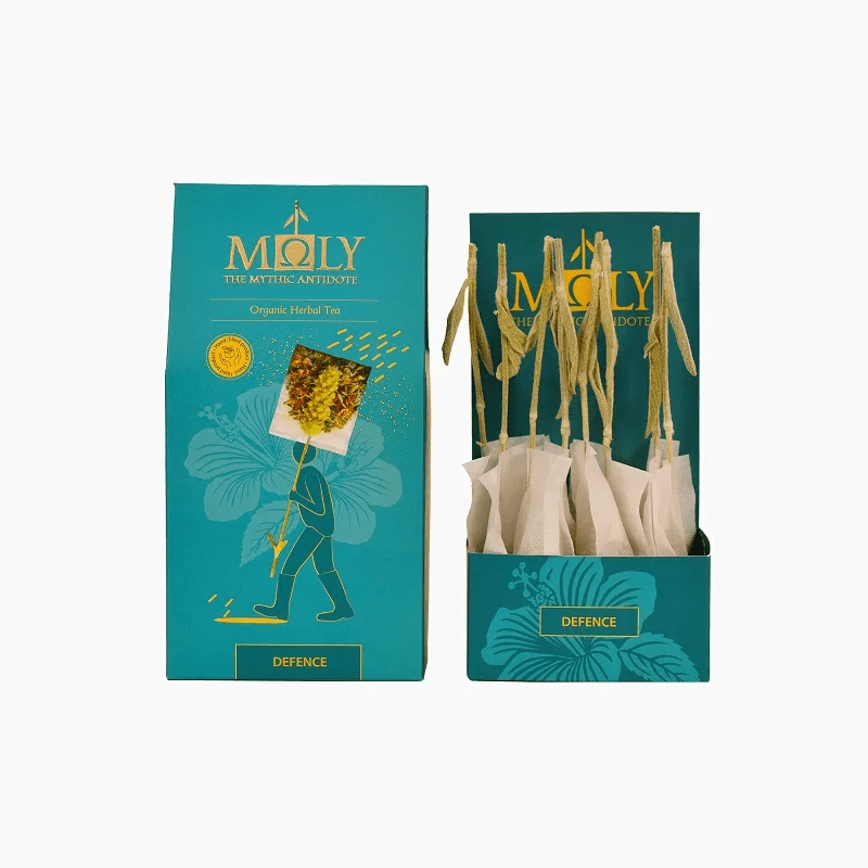 Teal retail box for Moly "Defence" Organic Herbal Tea shown next to an open display box containing individually wrapped tea bags with natural herbal stem stirrers.